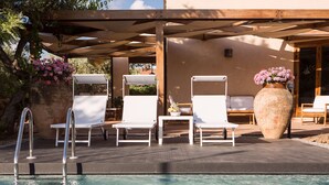 Zona de piscina con sillones de descanso, un jarrón grande con flores y una pérgola que ofrece sombra.