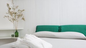 Una cama bien hecha con ropa blanca y una almohada verde, un florero verde con plantas secas sobre un estante blanco, y otra almohada blanca sobre la cama.