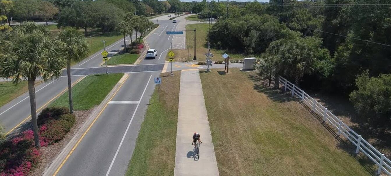 Un chemin pavé longe une route, où l’on aperçoit un cycliste, des palmiers et une clôture blanche.