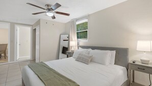 A hotel room with a bed, a nightstand, a lamp, a ceiling fan, a window with curtains, and a door leading to another room.