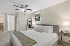 Une chambre d’hôtel comprenant un lit, une table de chevet, une lampe, un ventilateur de plafond, une fenêtre avec des rideaux et une porte donnant sur une autre pièce.