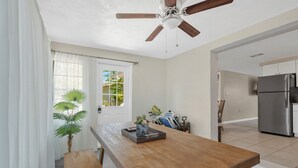 A dining area with a wooden table, benches, a ceiling fan, a door with a view, and a kitchen with a refrigerator.