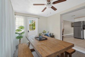 Un coin repas avec une table en bois, des bancs, un ventilateur de plafond, une porte donnant sur une vue agréable et une cuisine équipée d’un réfrigérateur.