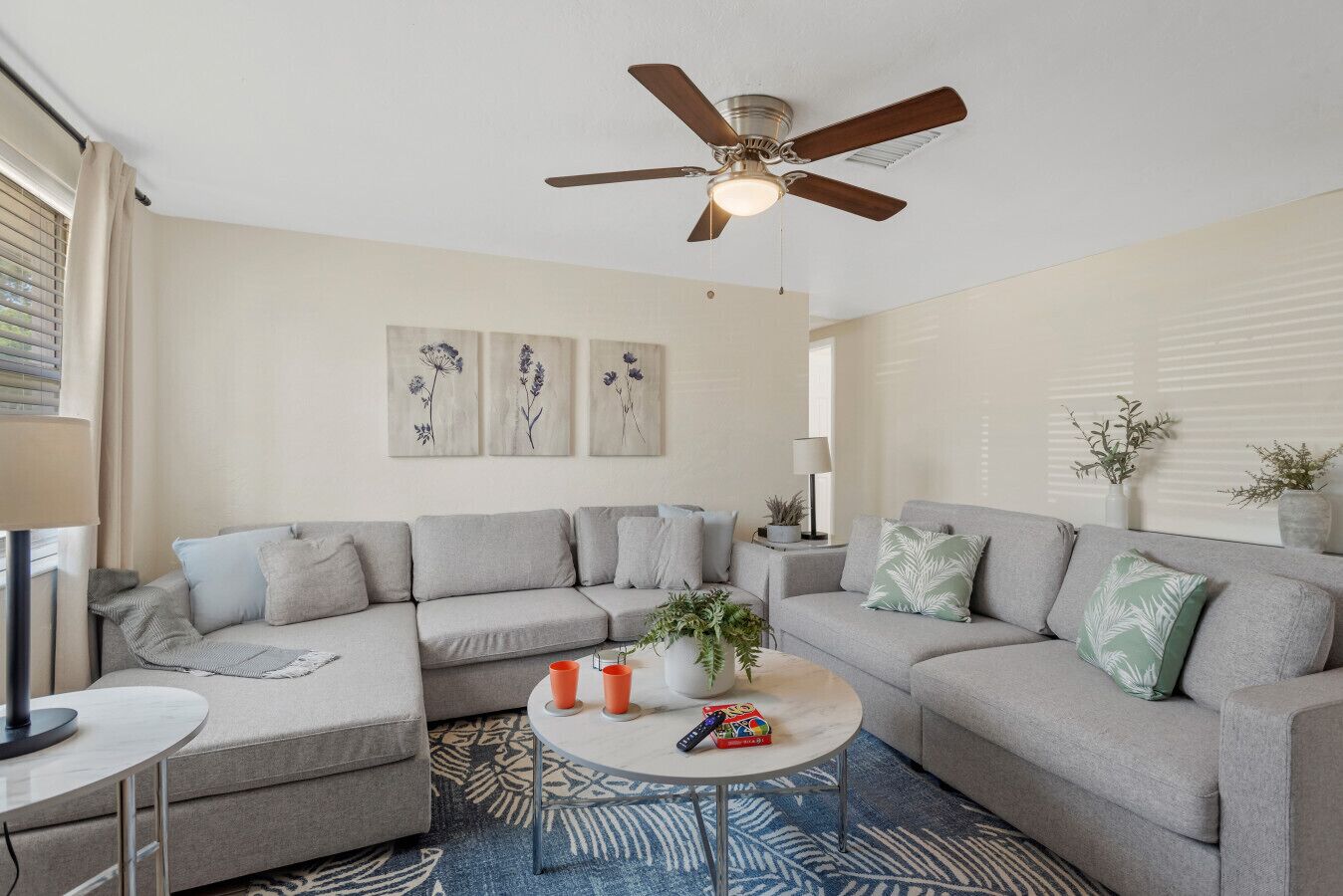 A living room with a sectional sofa, a ceiling fan, a coffee table with remote controls, and wall art.