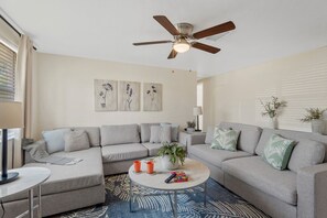 A living room with a sectional sofa, a ceiling fan, a coffee table with remote controls, and wall art.