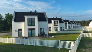 Eine Reihe moderner, weißer, eingeschossiger Häuser mit dunklen Dächern, jedes mit einer Terrasse und Gartenmöbeln.