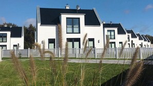 A row of modern, white houses with black roofs, white fences, and grassy areas.