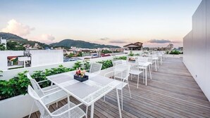 A rooftop terrace with white metal furniture and a wooden deck, overlooking a cityscape and hills. - LullaBella Hotel Patong (Patong)