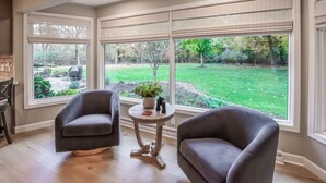 Una habitación con dos sillones oscuros, una mesita redonda con una planta y unos binoculares, y ventanas amplias que dan a un jardín cubierto de césped.
