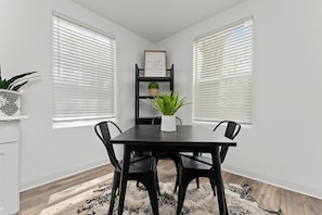 Ein Essbereich mit einem schwarzen Tisch, vier Stühlen, einem weißen Blumentopf, einem schwarzen Bücherregal und zwei Fenstern mit Jalousien.