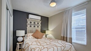 Ein Schlafzimmer mit einem Bett, Nachttischen, einer Wand-Klimaanlage und einem Fenster mit Jalousien.