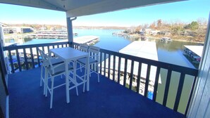 A balcony with a table and chairs, overlooking a calm lake with docks and buildings in the background. - Lakefront Corner Condo with Boat Slip - Stunning Views - Pool - Laundry (Osage Beach)
