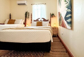 Une chambre d’hôtel avec deux lits, un sol carrelé avec un motif en briques, une fenêtre avec des rideaux légers et un climatiseur fixé au mur.