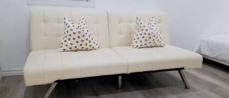 Un sofá de color crema con respaldo capitonado y patas metálicas, acompañado por dos almohadones de fondo blanco y lunares marrones.