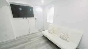 Une pièce avec un canapé blanc, un téléviseur à écran plat fixé au mur, une porte blanche et une fenêtre avec des stores.