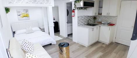Et lille hotelværelse med seng, et lille natbord, en hvid sofa og et tekøkken med hvide skabe og en vask.