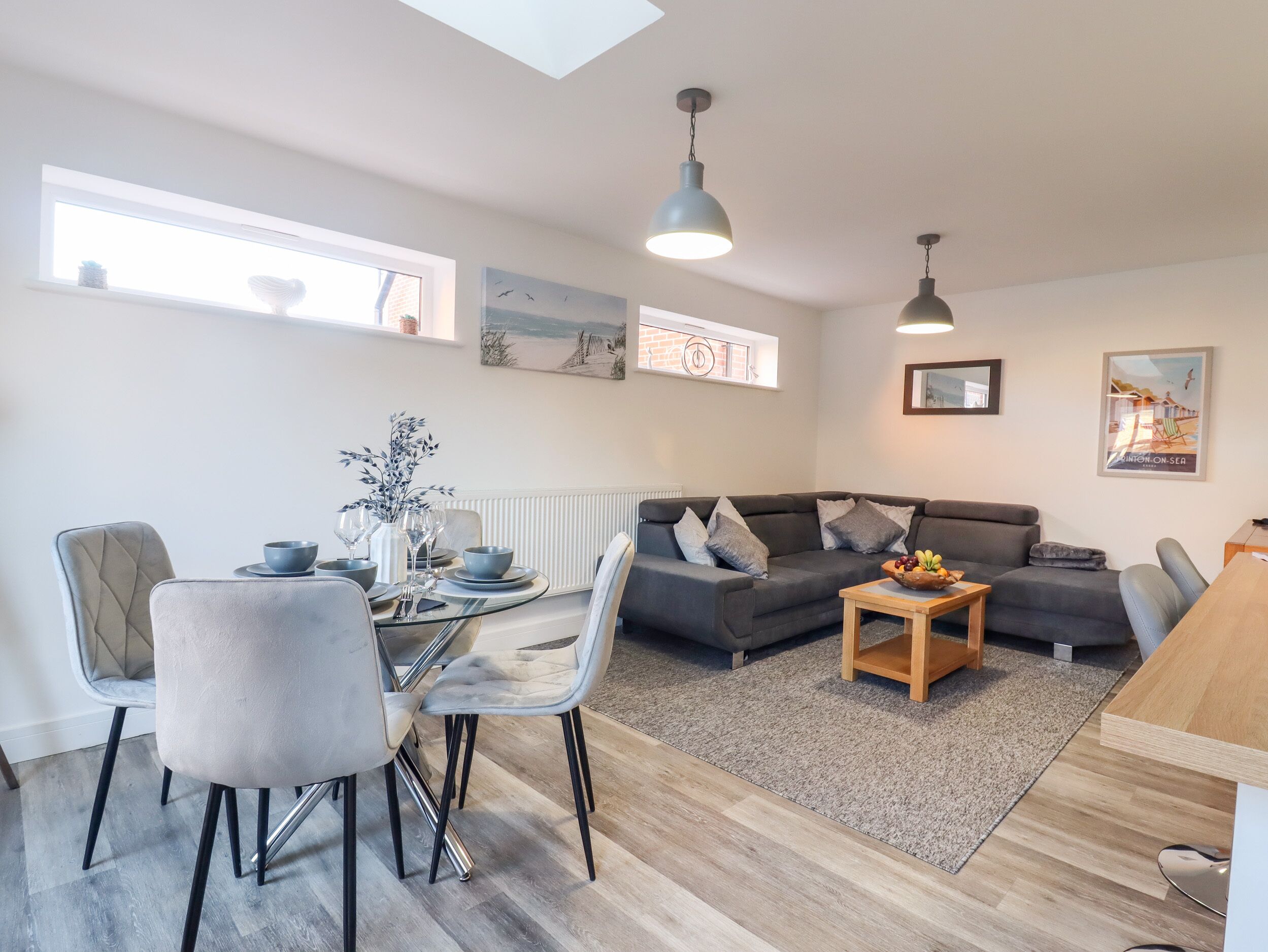 A modern living room with a dining area, grey sofas, a wooden coffee table, and a view of the sea through a window.