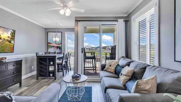 A modern living room with a grey sofa, a coffee table, and a dining area with a table and chairs. There is a large window with blinds, and a view of the outdoors. - Sands Beach Club | Ocean & Pool View | Amenities! (Myrtle Beach)