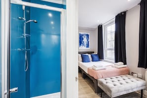 A modern hotel room with a blue shower enclosure, a bed with a red and white patterned bedspread, two blue pillows, a white bench, and a large window with black curtains. - Quality Spitalfields Accommodations (London)