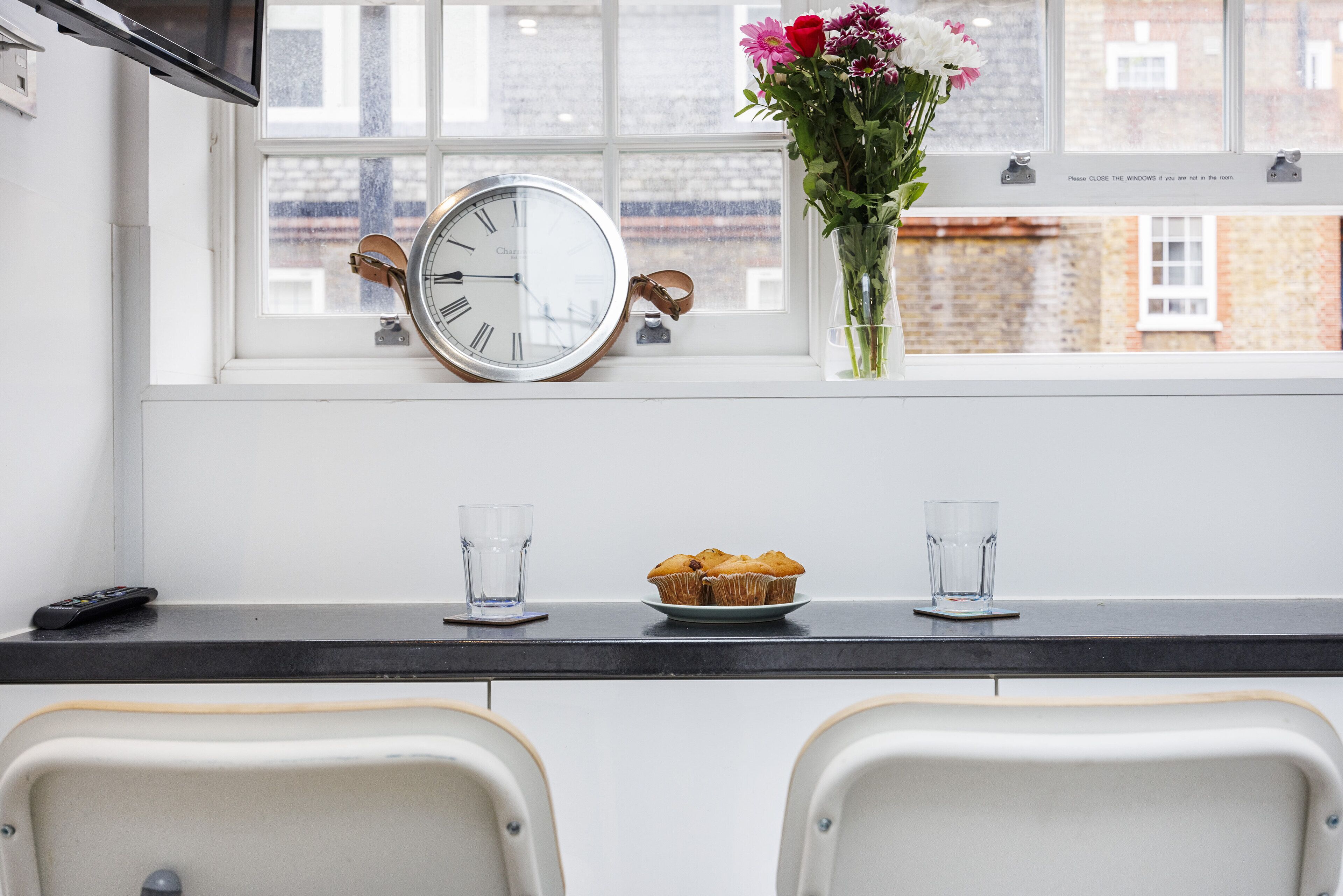 A kitchen with a clock, a vase of flowers, and a plate of muffins.
