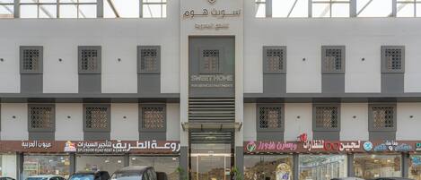 A multi-story building with a central entrance, several shops on the ground floor, and parked cars in front.