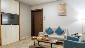 A living room with a flat-screen TV mounted on the wall, a wooden coffee table with a tray of food, a sofa with blue cushions, and a blue armchair. - Sweet Home Serviced Apartments (Jeddah)
