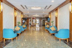 Un hall d’hôtel avec un sol en marbre, des fauteuils bleus aux pieds en bois et un comptoir d’accueil central.