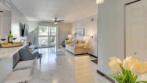 A modern living room with a sofa, a dining area with a table and chairs, a kitchen area with a counter and chairs, and a large window providing natural light. - Summer Getaway: Destin Condo by Golf & Dining (Destin)