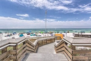 Ein hölzerner Steg führt zu einem Strand mit Sonnenschirmen und Menschen.