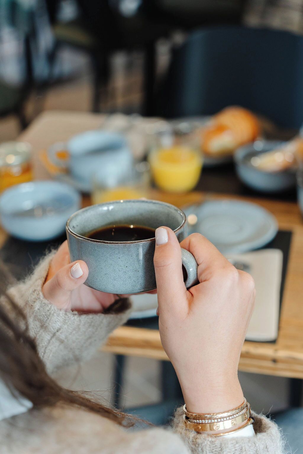 A person holding a ceramic mug with a dark liquid, likely coffee, in a casual dining setting with various dishes and a laptop on the table.