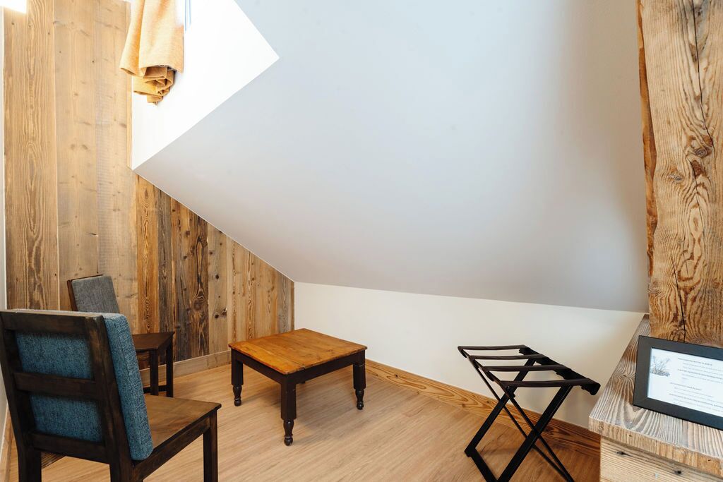 A room with a wooden floor, a small wooden table, a blue chair, a black folding chair, and a white wall with a wooden panel.
