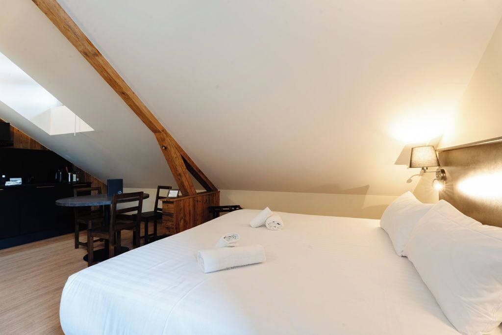 A bedroom with a sloped ceiling, a bed with white linens, a desk with a chair, and a lamp on the wall.