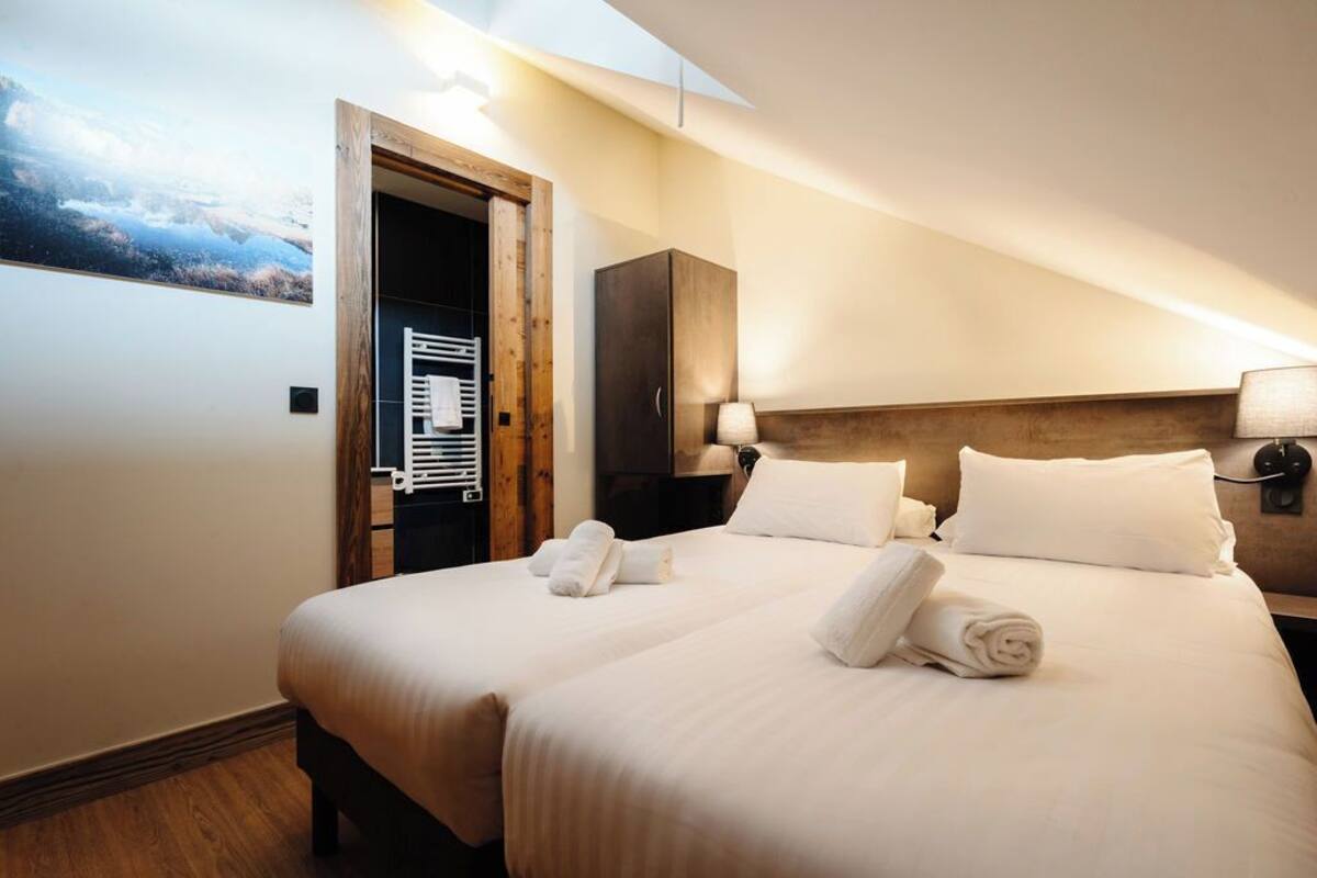 A hotel room with a large bed, two bedside tables with lamps, a wooden headboard, and a view of a mountainous landscape through an open door.