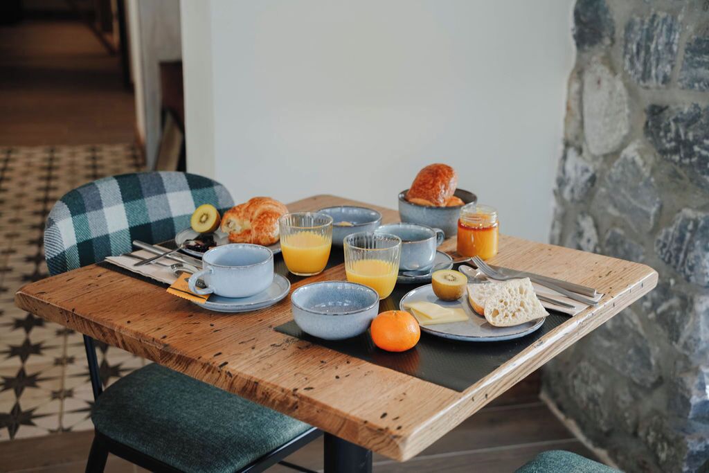 Un desayuno con una mesa de madera, una silla a cuadros y un muro de piedra al fondo.