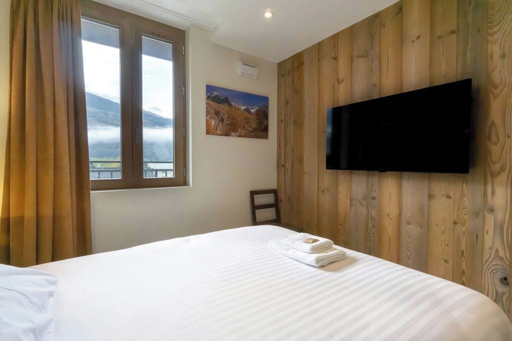 A hotel room with a bed, a flat-screen TV mounted on the wall, a window with curtains, and a painting on the wall.