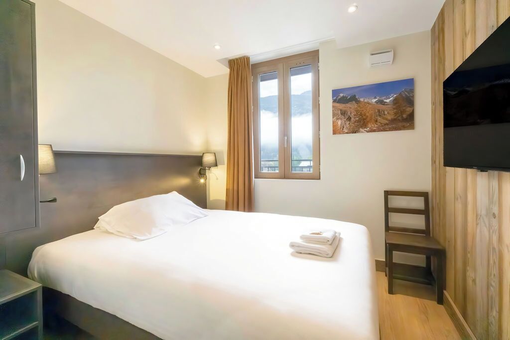 A hotel room with a bed, a nightstand, a window with curtains, a wall-mounted TV, and a painting.