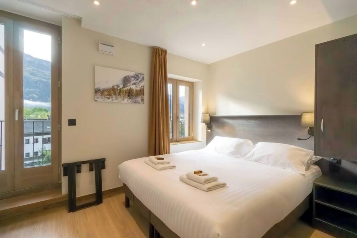 A hotel room with a large bed, a view of mountains through the window, a painting on the wall, and a TV on the side.
