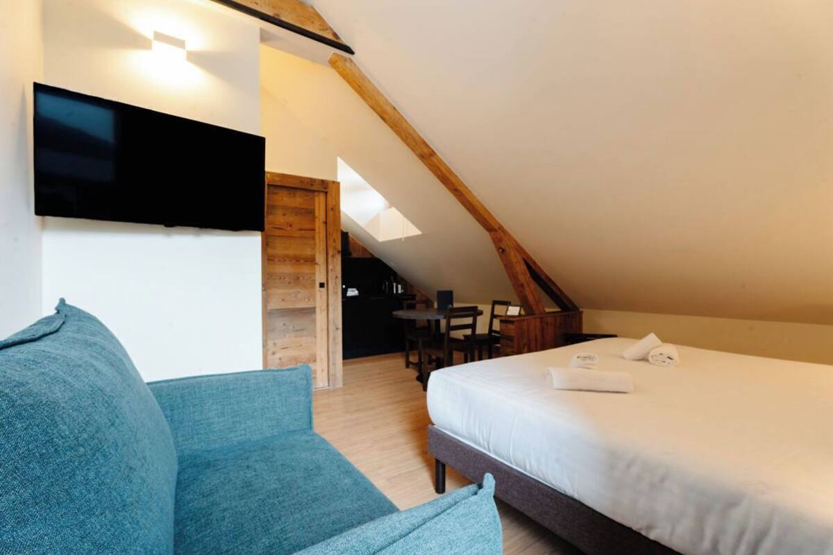 A modern room with a bed, a sofa, a TV mounted on the wall, and a dining area with a table and chairs.