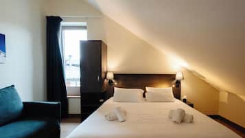 Una camera d'albergo con un letto, due lampade ai lati, una testiera scura, una finestra con tende, un divano scuro e un comodino con lampada.