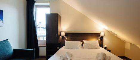 Habitación de hotel con cama, dos lámparas de noche, cabecera oscura, ventana con cortinas, sofá oscuro y mesita de noche con lámpara.