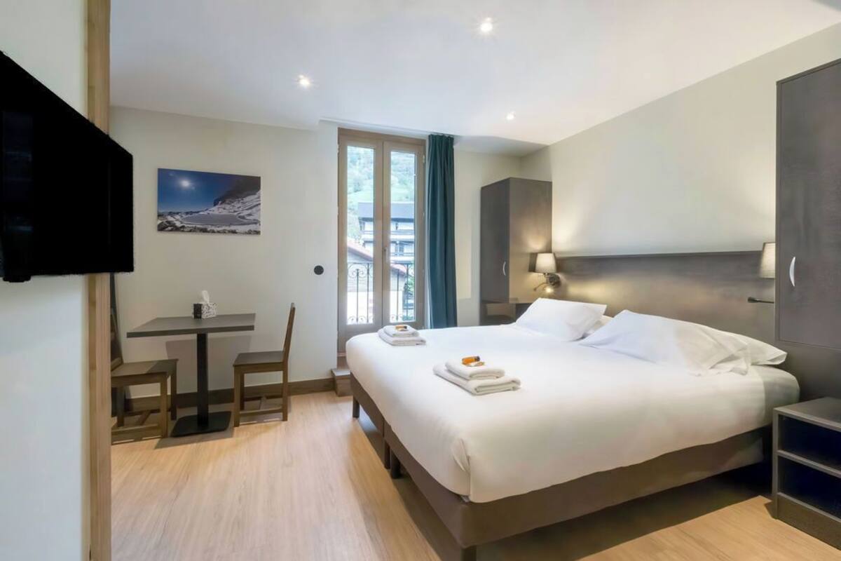 A hotel room with a bed, a desk, a chair, a TV, and a window with a view of the outside.