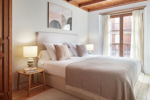 A bedroom with a large bed, two bedside lamps, a small table with a book, and a window with curtains.