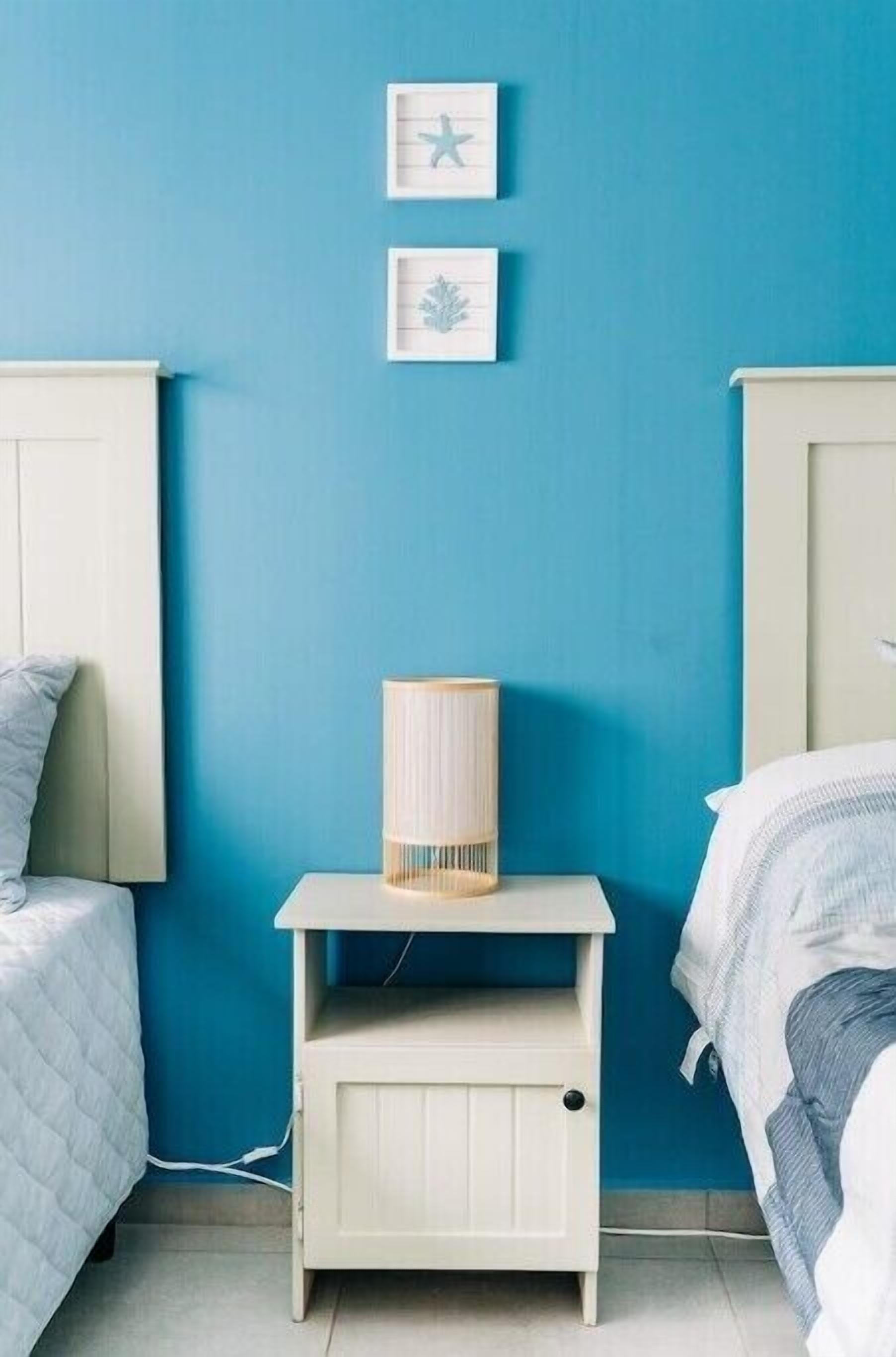 A bedroom with a blue wall, a bedside table with a lamp, and two framed pictures.