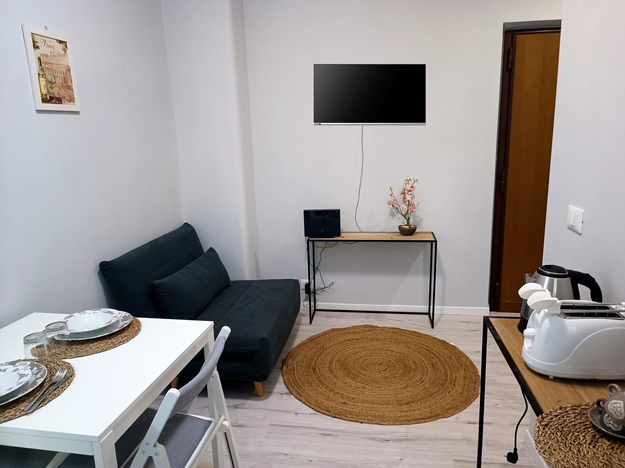 Un ambiente compatto con divano, un tavolo da pranzo per due persone, una televisione a schermo piatto montata a parete, un piccolo tavolino con un vaso di fiori e una macchina da caffè.