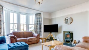 A living room with a brown leather sofa, a blue ottoman, a wooden coffee table, a flat-screen TV, and a fireplace. - Tre Lowen (Port Isaac)