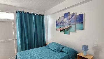 Un dormitorio con una cama, cortinas verde mar, una mesita de noche de madera, una lámpara y tres cuadros en la pared.