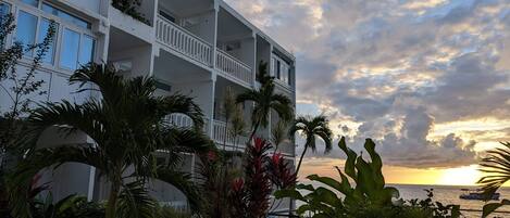 Un hotel de varios pisos con balcones, rodeado de plantas tropicales y una vista del atardecer sobre el mar.