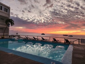 Una piscina con sillas de descanso, vista al océano y un atardecer.