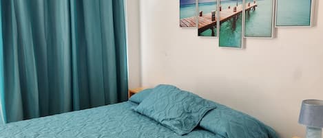 Une chambre à coucher avec un lit, des rideaux de couleur turquoise, une table de chevet en bois et un mur orné de quatre tableaux encadrés.