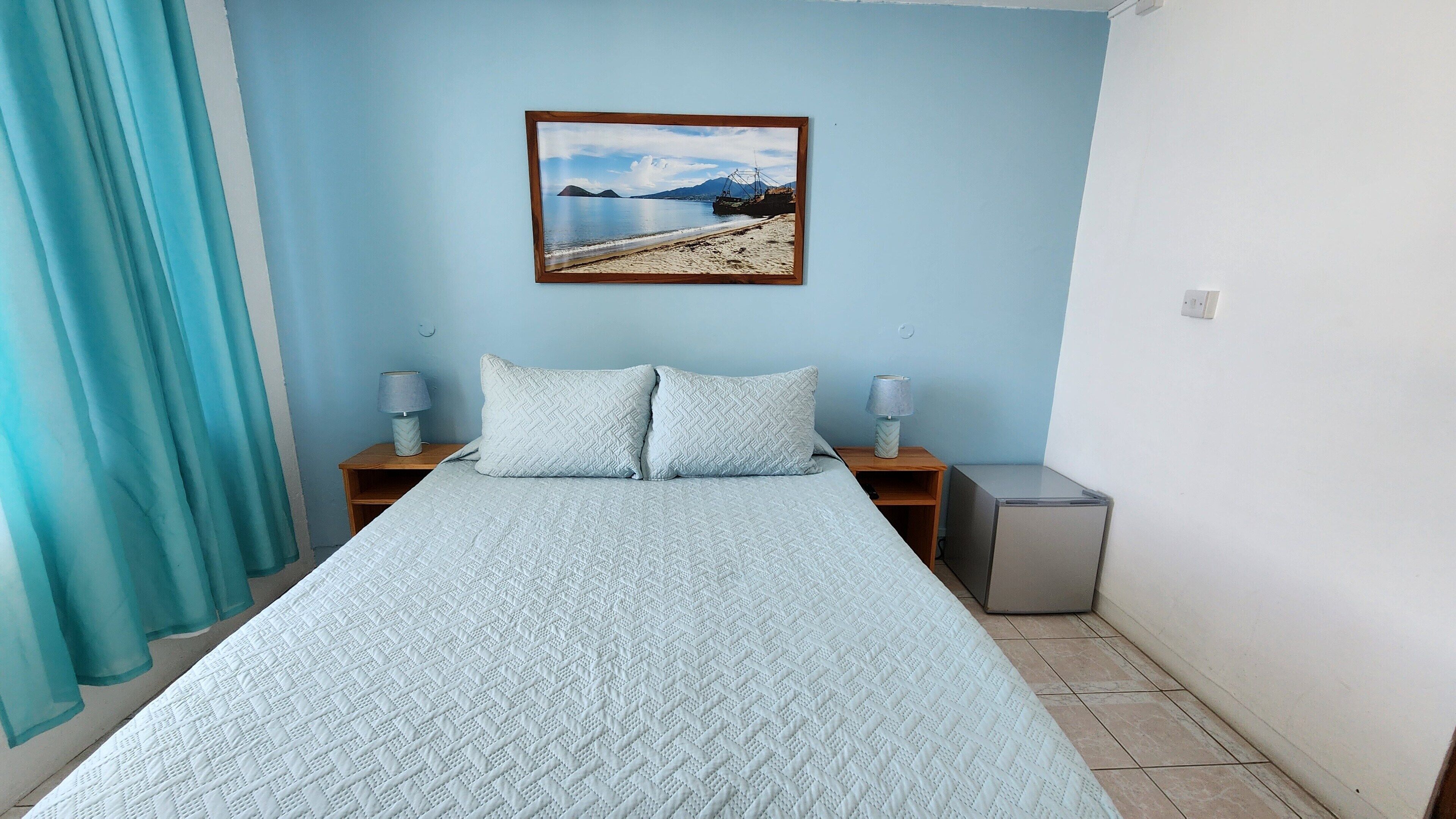 A bedroom with a bed, two nightstands, a lamp, a picture on the wall, and a small refrigerator.